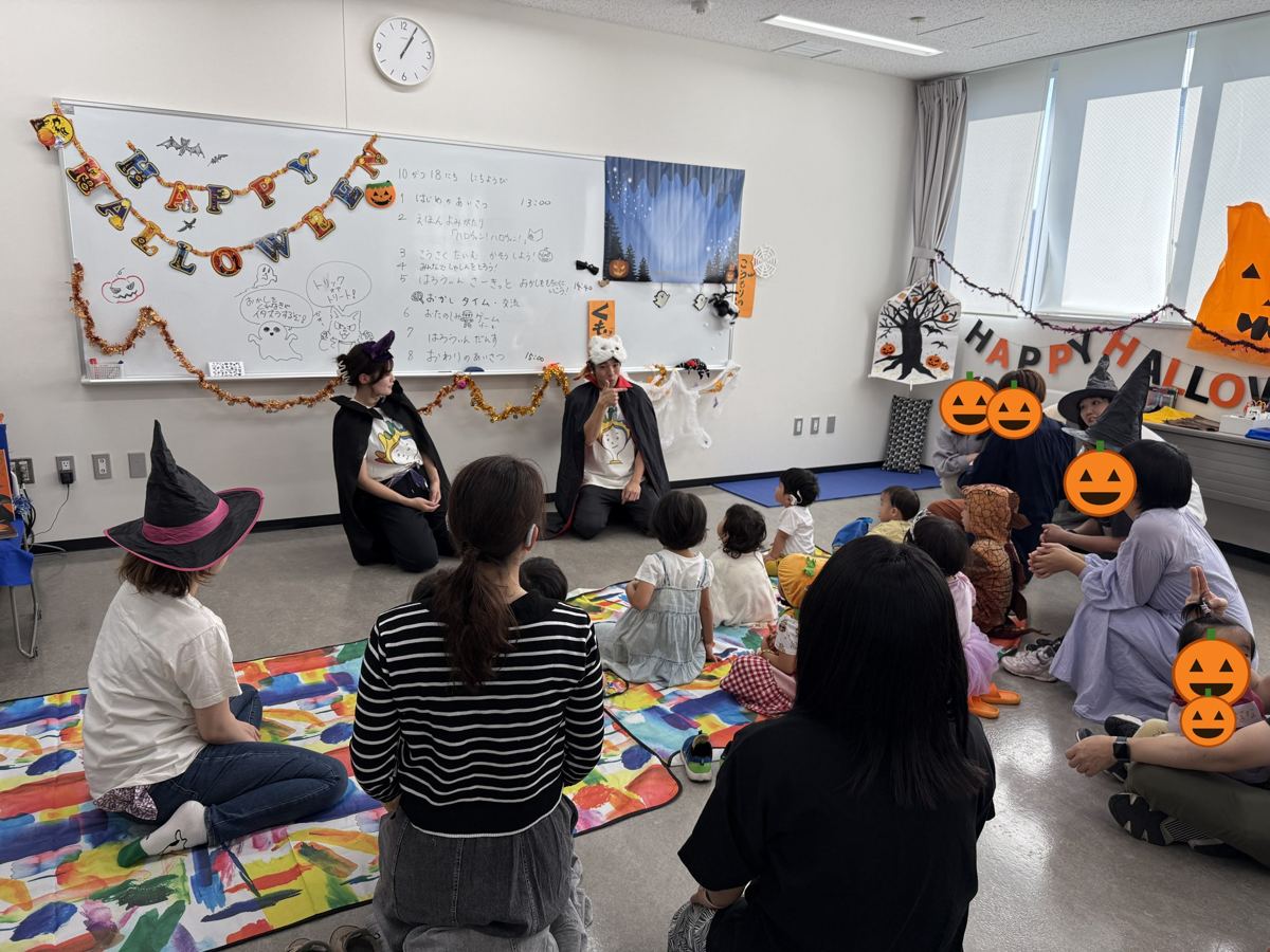 ハロウィンの飾りつけの部屋で、手話で絵本を読み語るスタッフと子どもたち